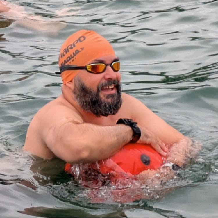 Francis Szakacs in the water in an orange swim cap and mirrored goggles, holding an orange tow float
